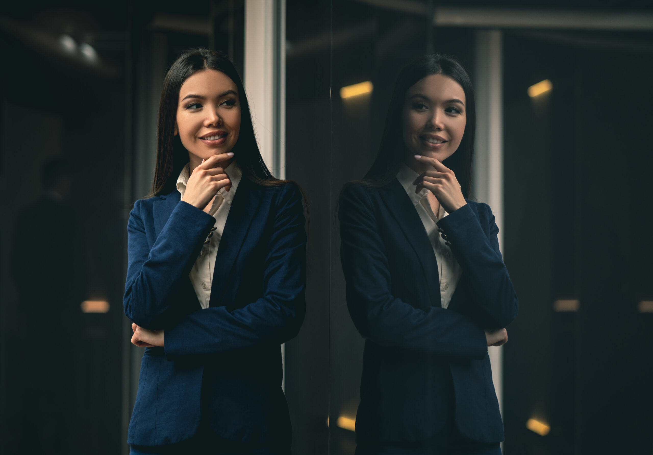 A professional skilled woman captured in a moment of reflection, standing in front of a glass wall in an office environment.